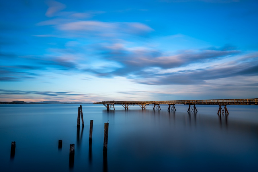 The view of the ocean with still water, a long pier and clouds above.